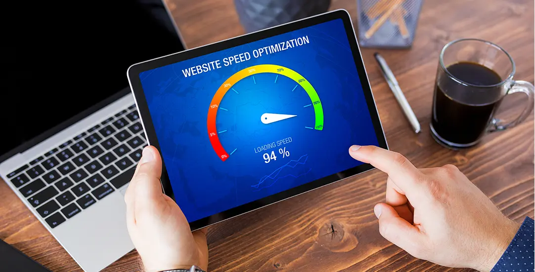 Person holding a tablet displaying a “Website Speed Optimization” dashboard with a gauge showing 94% loading speed, next to a laptop and a cup of coffee on a wooden desk.