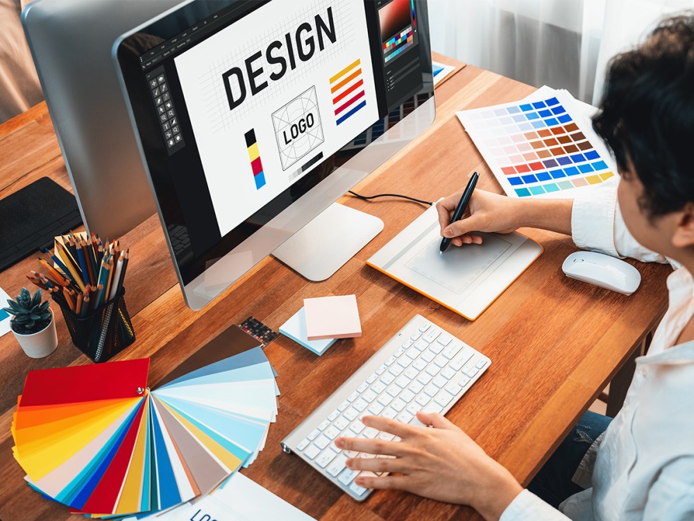 A person working at a desk using a graphics tablet and computer to design a logo, with color swatches, pencils, and design tools spread out around the workspace. - Shark Web A person working at a desk using a graphics tablet and computer to design a logo, with color swatches, pencils, and design tools spread out around the workspace.